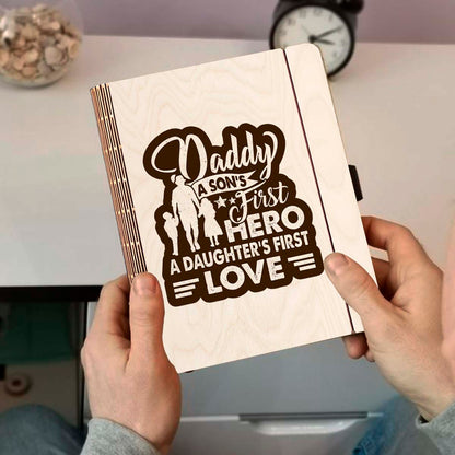 Detail crop on the wooden notebook cover, with father-and-children silhouette plus 'Daddy first hero /, sharp birch grain.